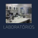 A imagem mostra uma fotografia de um laboratório emoldurada por um fundo azul marinho.Podemos ver um ambiente interno com bancadas brancas e equipamentos de laboratório. Há microscópios, frascos, computadores e outros aparelhos. Ao fundo, existem armários brancos com portas de vidro, contendo mais equipamentos e materiais. Abaixo da fotografia, em letras brancas maiúsculas e grandes, está escrito "LABORATÓRIOS".