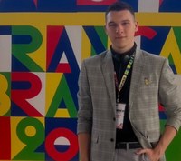 Homem jovem em pé diante de um painel colorido com letras geométricas. Ele veste paletó cinza xadrez sobre camisa preta e crachá de evento pendurado no pescoço. A composição do fundo possui cores vivas — vermelho, azul, verde, amarelo e branco — em estilo gráfico moderno.