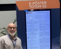 Homem de barba grisalha e óculos sorri. Na tela vertical do totem é exibido um pôster eletrônico com texto acadêmico e logotipo da ABRASCÃO no topo. O ambiente é interno e iluminado, com plantas ao fundo e atmosfera de evento científico.